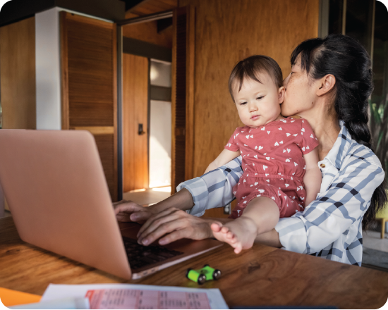 A parent working at a laptop while holding a young child